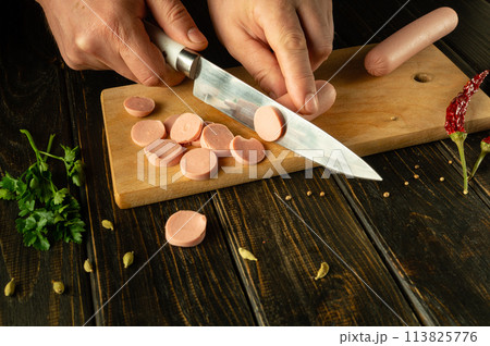 A man cuts vienna sausage on a wooden cutting board. Preparing delicious sandwiches for dinner on the kitchen table at home 113825776