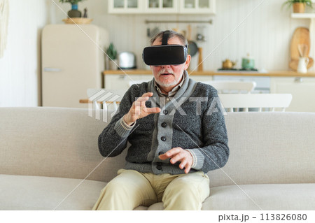 Excited mature senior man wearing using virtual reality metaverse VR glasses headset at home. Grandfather touching air during VR experience on virtual reality helmet. Simulation hi-tech videogame 113826080