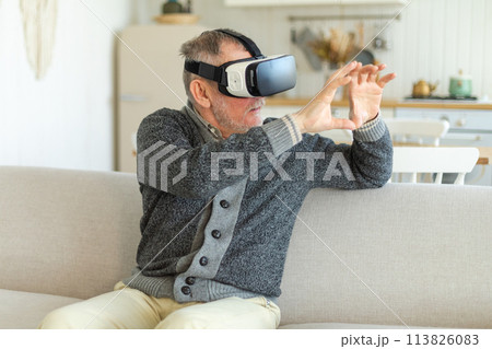 Excited mature senior man wearing using virtual reality metaverse VR glasses headset at home. Grandfather touching air during VR experience on virtual reality helmet. Simulation hi-tech videogame 113826083