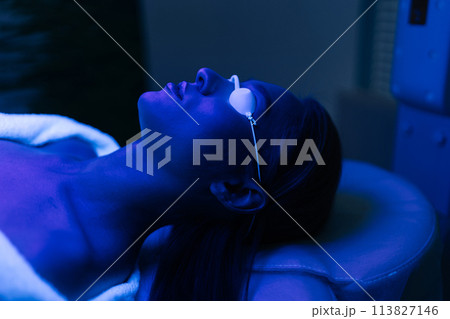Closeup face of female patient undergoes blue LED light therapy for skin rejuvenation at modern aesthetic clinic. Using phototherapy device with blue light wavelength for acne treatments. Closeup face of female patient undergoes blue LED light therapy for skin rejuvenation at modern aesthetic clinic. Using phototherapy device with blue light wavelength for acne treatments. 113827146