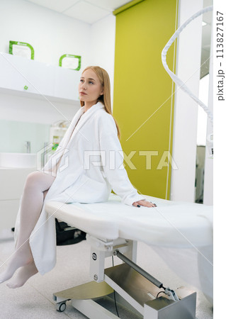 Vertical shot of pretty blonde female patient in white bath robe sitting on medical couch in modern cosmetology clinic while waiting beautician specialist. Concept of cosmetology, beauty treatment 113827167