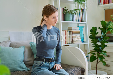 Young teenage female experiencing neck pain sitting on couch at home Young teenage female experiencing neck pain sitting on couch at home 113829862