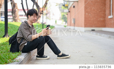 A happy Asian male college student sits on the street in a campus, using his smartphone. 113835736