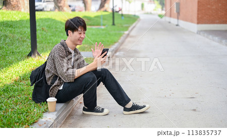 A cheerful Asian student sits on the street talking with his friend on a video call on his phone. 113835737