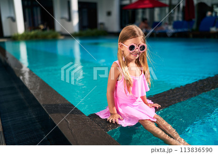 Little girl in pink dress and funny sunglasses sits by poolside. Child enjoys sunny day at outdoor swimming pool, summer fun for kids. Bright sunlit leisure moment, playful water activity. Little girl in pink dress and funny sunglasses sits by poolside. Child enjoys sunny day at outdoor swimming pool, summer fun for kids. Bright sunlit leisure moment, playful water activity. 113836590