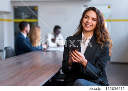 Confident businesswoman in modern office smiles, uses smartphone. Team collaborates in background. Professional woman in suit multitasks, engages in mobile tech communication, team strategy session. 113836615
