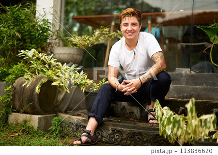 Smiling Gen Z Individual sits on steps, exhibits comfort, confidence in coworking space, exemplifying transgender pride in LGBTQ friendly workspace, reflects modern digital nomad lifestyle. 113836620