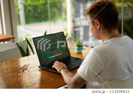 Focused crypto trader analyzes market data on laptop screen in modern coworking space. Person trading digital currency, studying graphs, trends. Freelancer in casual wear working remotely, investing. 113836632