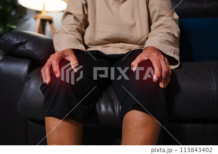 close up senior woman suffering from knee pain while sitting on sofa in living room at night 113843402
