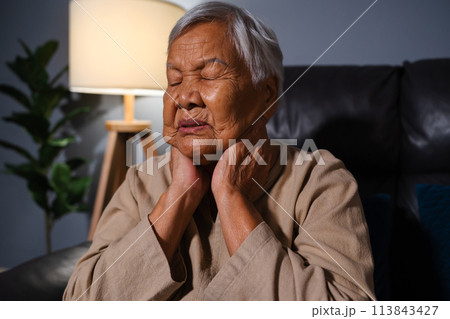 senior woman suffering from neck pain while sitting on sofa in living room at night 113843427
