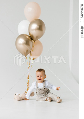 Birthday baby boy is sitting on the floor, with white, gold and beige balloons next to him Birthday baby boy is sitting on the floor, with white, gold and beige balloons next to him 113844678