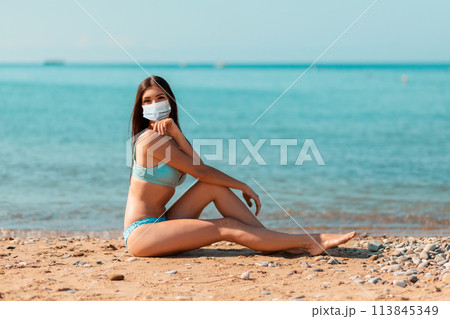 A young woman in a protective mask, sitting on the ocean and posing. The concept of home away from home during a pandemic coronavirus A young woman in a protective mask, sitting on the ocean and posing. The concept of home away from home during a pandemic coronavirus 113845349