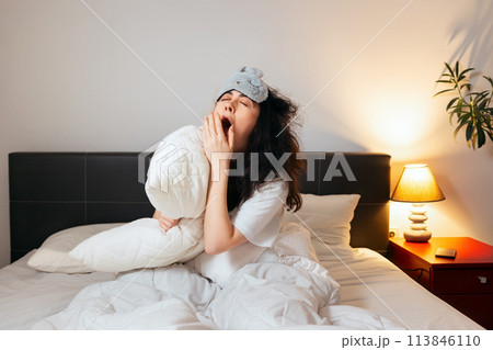 Tired Caucasian woman wearing sleep mask sitting in bed with pillow and very yawning. Concept of insomnia and sleepless 113846110