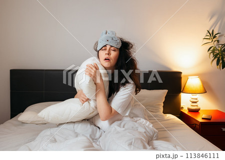 Tired Caucasian woman wearing sleep mask sitting in bed with pillow and can't sleep. Concept of insomnia and sleepless 113846111