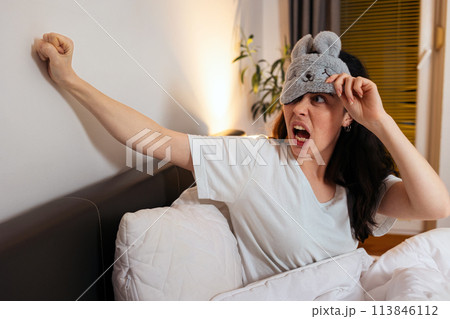 Portrait of annoyance angry Caucasian woman removing sleep mask knocking with fists to wall sitting in bed. Insomnia due to noisy neighbors Portrait of annoyance angry Caucasian woman removing sleep mask knocking with fists to wall sitting in bed. Insomnia due to noisy neighbors 113846112