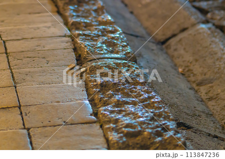 A stone walkway with a nearby passing groove for draining water. Evening light reflections on various objects 113847236