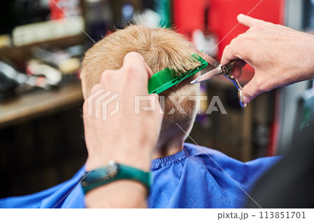 Man barber cutting little boy's hair using comb and scissors. Child getting haircut from adult male, likely barber. Professional hairdresser and cute client at modern barbershop. 113851701