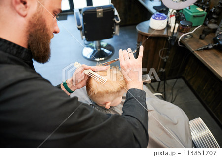 Professional hairdresser making stylish haircut for little client. Behind shoulder view of man cutting hair. Barbershop worker making short men's haircut. 113851707