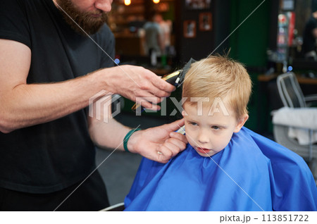 Hairdresser using electric shaver to cut boy's hair. Little kid getting first haircut in barbershop. Close up Hairdresser using electric shaver to cut boy's hair. Little kid getting first haircut in barbershop. Close up 113851722