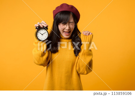 A young Asian woman with a happy smile, wearing a yellow sweater and red beret, stands confidently next to an alarm clock in her stylish office. 113859106