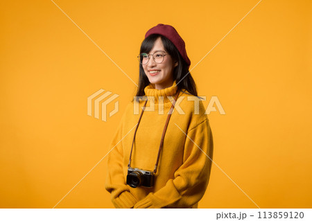 With her vintage camera in hand, a young Asian woman, donning a yellow sweater and red beret, pursues her passion for photography against a vibrant yellow background. With her vintage camera in hand, a young Asian woman, donning a yellow sweater and red beret, pursues her passion for photography against a vibrant yellow background. 113859120