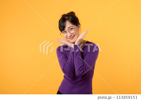 A shy and cute young woman in her 30s wearing purple shirt and eyeglasses, expressing happiness with a joyful smile. Positive emotions in a yellow studio background. A shy and cute young woman in her 30s wearing purple shirt and eyeglasses, expressing happiness with a joyful smile. Positive emotions in a yellow studio background. 113859151