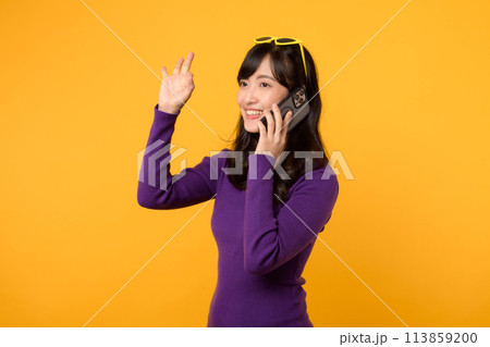 Radiate joy with a young Asian woman in her 30s, wearing an purple shirt, showing okay sign gesture while using smartphone on yellow background. 113859200