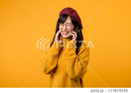 Chic girl in a vibrant beret and yellow sweater, sharing a happy moment on her smartphone against a cheerful yellow background. 113859248