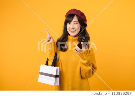 Young Asian woman in her 30s, holding a shopping paper bag and smartphone, showcasing her cheerful demeanor in a yellow sweater and red beret against a yellow background, making an online purchase. 113859297