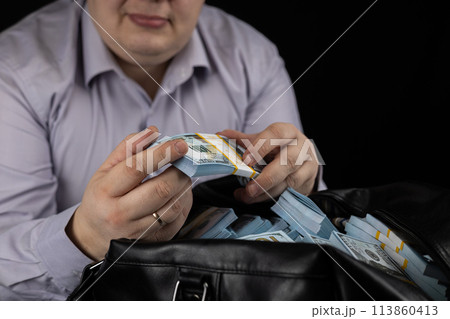 A man runs his finger over a stack of dollars in his hand, flipping through them on a black background. A man flips through dollars over a large bag of money. Unexpected big earnings concept. 113860413
