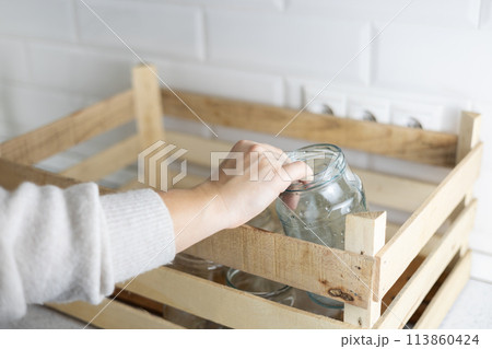 Glass recycling. A woman's hand puts a glass jar into a drawer on the kitchen table. The wooden box already contains a lot of old glass jars for recycling. Zero waste concept. 113860424