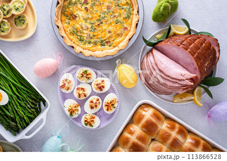 Easter brunch on large table with spiral sliced ham, quiche, deviled eggs and hot cross buns 113866528