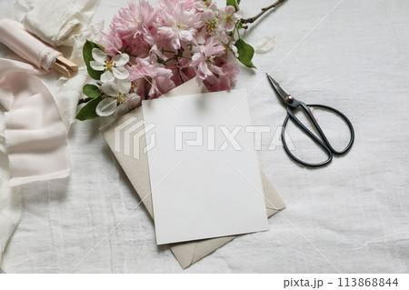 Wedding spring styled photo. Feminine scene mockup with pink blossoming Japanese cherry, apple tree branch. Blank paper greeting card, black scissors, white linen table background. Flat lay, top view. 113868844