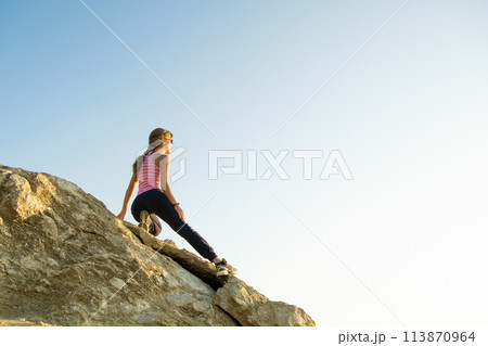 Woman hiker climbing steep big rock on a sunny day. Young female climber overcomes difficult climbing route. Active recreation in nature concept. 113870964