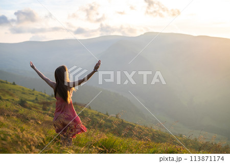 Back view of young happy woman traveler in red dress standing on grassy hillside on a windy evening in summer mountains with outstretched arms enjoying view of nature at sunset. 113871174
