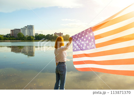 Young womanl with USA national flag on her shoulders with high city buildings in background celebrating United States independence day. 113871370