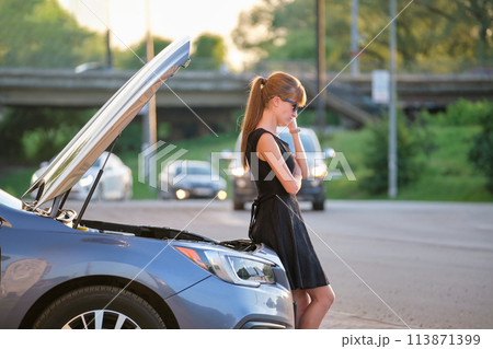 Sad female driver standing on a city street near her car with popped up hood and broken engine waiting for road service assistance to arrive 113871399