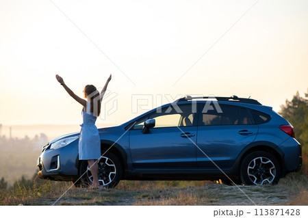 Happy young woman driver in blue dress enjoying warm summer evening standing beside her car. Travelling and vacation concept. Happy young woman driver in blue dress enjoying warm summer evening standing beside her car. Travelling and vacation concept. 113871428
