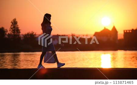Lonely pedestrian woman walking alone on lake side on warm evening. Solitude and relaxation concept. 113871465