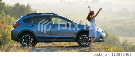 Young female driver resting near her car raising up her hands on warm summer evening. Travel destinations concept. 113871468