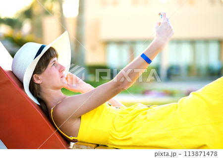 Young woman in yellow summer dress taking selfie with her mobile phone resting on a bench in park. Young woman in yellow summer dress taking selfie with her mobile phone resting on a bench in park. 113871478