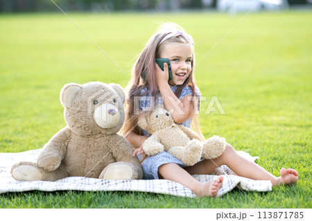 Pretty child girl sitting in summer park on green grass lawn with her teddy bear toy talking on mobile phone outdoors in summer. Pretty child girl sitting in summer park on green grass lawn with her teddy bear toy talking on mobile phone outdoors in summer. 113871785