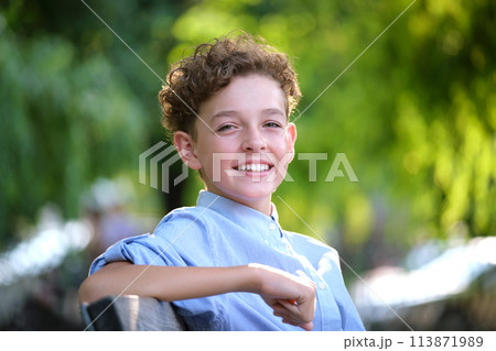 Young happy child boy relaxing sitting on bench in summer park. Positive kid enjoying summertime outdoors. Child wellbeing concept 113871989