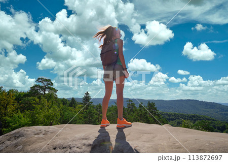 Sportive woman standing alone on hillside trail. Female hiker enjoying view of summer nature from rocky cliff on wilderness path. Active lifestyle concept 113872697