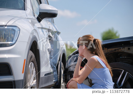 Sad young woman driver sitting near her smashed car looking shocked on crashed vehicles in road accident Sad young woman driver sitting near her smashed car looking shocked on crashed vehicles in road accident 113872778