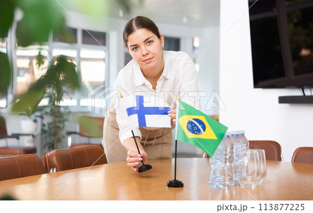 Young female assistant putting up brazilian and finnish flags on table for international negotiations 113877225