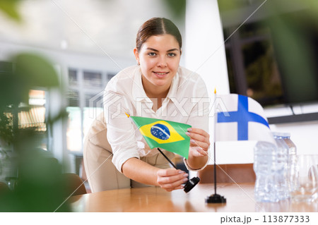 Positive young woman putting flag of Brazil on the table with flag of Finland in conference room Positive young woman putting flag of Brazil on the table with flag of Finland in conference room 113877333