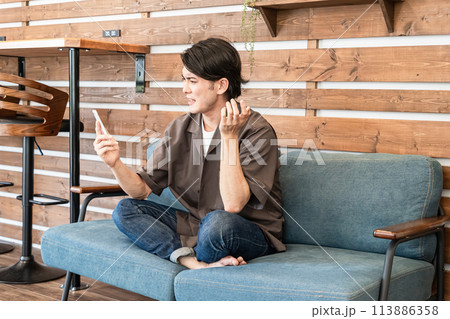 家でくつろぎながらスマホ・スマートフォンをいじる若い男性（困る・返信・メール・悩む・迷う） 113886358