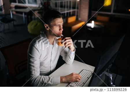 Pensive handsome programmer eating burger working in office at night. Closeup of professional male employee sitting on table, feeling hungry and starving, having dinner in corporate workplace 113899226