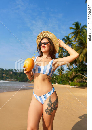 Sunlit young woman in stylish bikini holds fresh coconut on sandy beach. Fit body, traveler enjoys tropical vacation. Blue skies, palm trees background with copy space. Summer getaway, leisure moment. Sunlit young woman in stylish bikini holds fresh coconut on sandy beach. Fit body, traveler enjoys tropical vacation. Blue skies, palm trees background with copy space. Summer getaway, leisure moment. 113900863